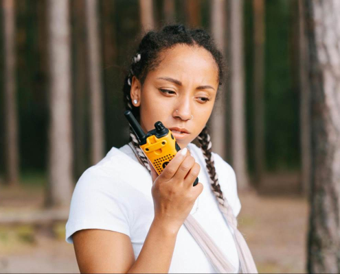 A woman standing in a forested area trying to communicate using a two-way radio