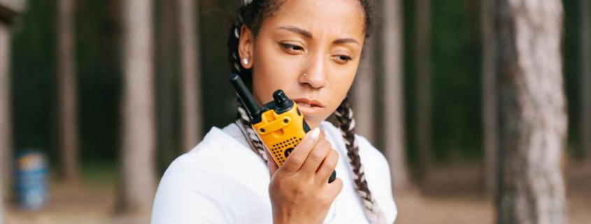 A woman standing in a forested area trying to communicate using a two-way radio