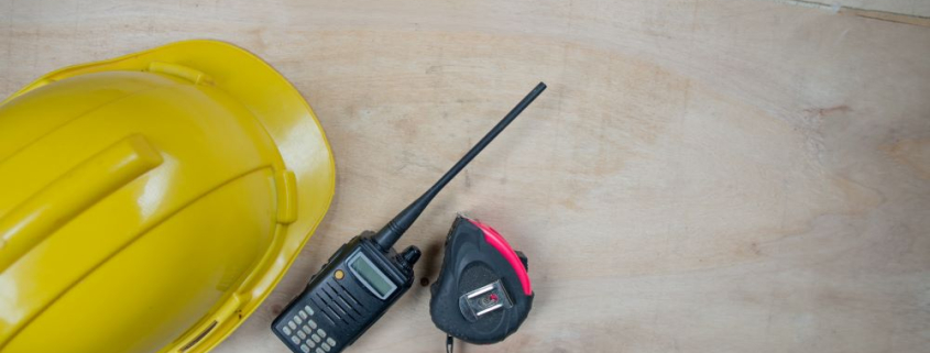 A portable black two-way radio is kept on a table-top, beside a helmet